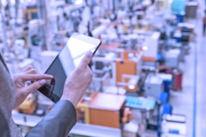 Horizontal color image of businesswoman - unrecognizable person - working with digital tablet in large futuristic factory. Woman standing on top of a balcony, holding touchpad and checking inventory of a manufacturing company on touchscreen tablet. Focus on businesswoman's hands holding black tablet, futuristic machines in background.